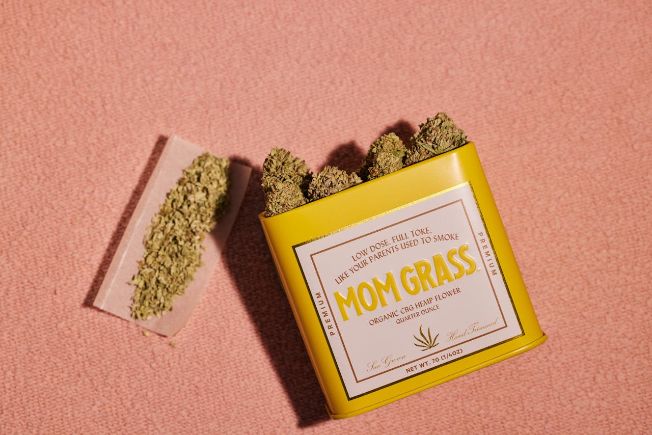 Yellow tin labeled 'Mom Grass' with hemp buds and rolled hemp on soft background.